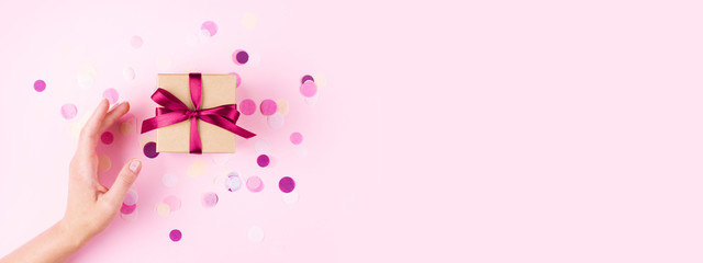 Woman hand and kraft gift box with pink bow on pink background decorated with confetti.. Top view, holiday present concept. Banner for site.