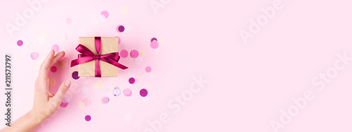 Woman hand and kraft gift box with pink bow on pink background decorated with confetti.. Top view, holiday present concept. Banner for site.