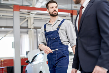 Portrait of modern bearded mechanic talking to unrecognizable businessman working across workshop in  car service and repair center, copy space