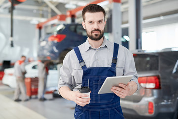 Waist up portrait of modern bearded mechanic holding digital tablet and cup of coffee  posing...