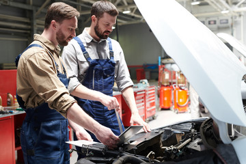 Portrait of two modern bearded mechanics looking under hood of car inspecting engine and using...