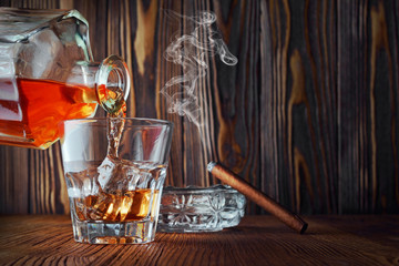Strong alcoholic drink scotch whisky pour from decanter in old fashion glass with ice cube and smoking cigar in ashtray