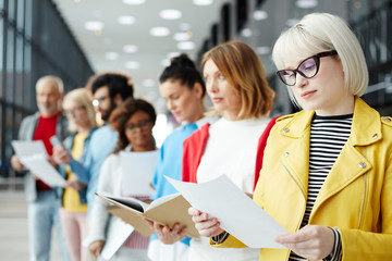 Blond young woman in eyeglasses reading her resume while standing in queue and waiting for her turn for interview