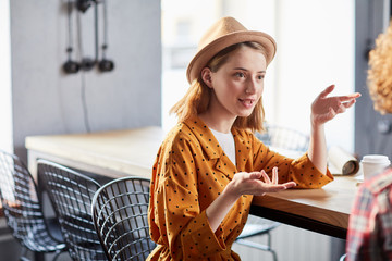 Young stylish businesswoman in hat discussing working points with her colleague in cafe