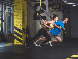 Full length side view beaming male and cheerful woman speaking while making workout with suspension cable in fitness center
