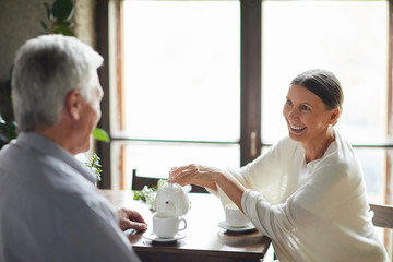 Smiling senior wife making tea for her husband while both sitting in cafe