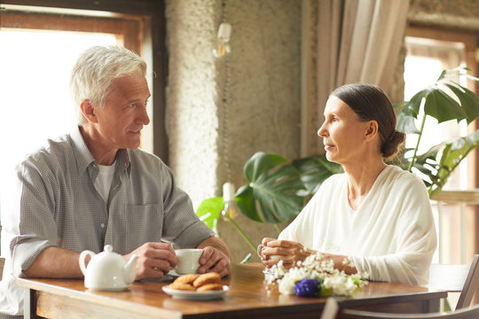 Restful senior couple sitting by table in cafe, having tea with cookies and talking