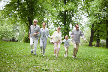 Obraz premium Cheerful and energetic family running on green grass among trees in summer park