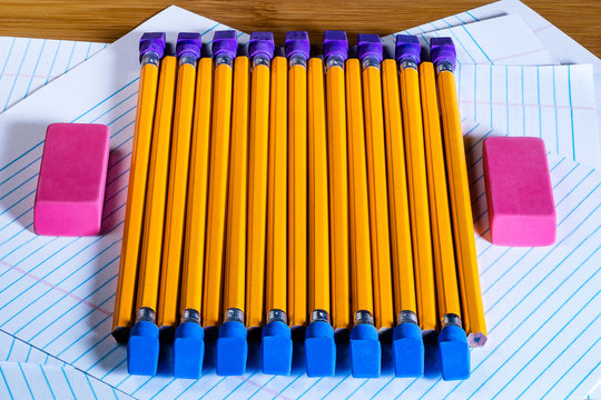 Several pencils in opposing direction with pencil erasers placed on one end arranged on a disarray of blank filler paper sheets; on either side sits a large eraser.