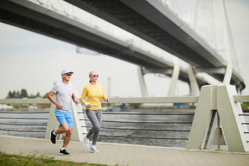 Contemporary senior couple in activewear jogging on bridge along waterside in the morning