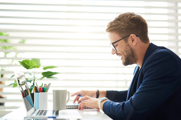 Profile of smiling male sitting at desktop and touching notebook. There are laptop, coffee mug and colorful markers on table