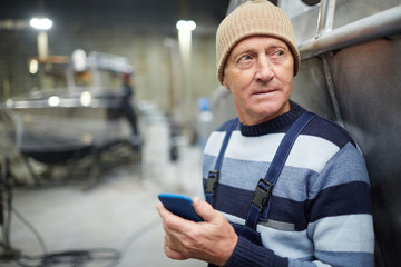Pensive senior technician in workwear texting in his smartphone while standing by ship