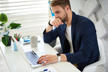 Side view of thoughtful male sitting at table with laptop. He is typing and touching mouth with one hand while making important decision at job. Coffee cup and notebook with pens and glasses are aside