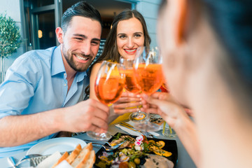 Handsome Middle-Eastern young man sitting next to his Caucasian girlfriend while toasting with two mutual friends outdoors at a trendy restaurant in summer