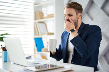 Fototapeta premium Sleepy morning. Side view of male holding cup of coffee and yawning. He is covering mouth with hand sitting at desktop with computer