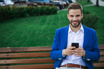 Portrait of happy man using cellphone while sitting on bench in park. He is looking at camera and smiling. Copy space 