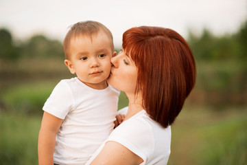 Mother and little son together outdoor. Mother holds her son in arms and tenderly kisses him. Green background. Happy family and mother love and care concept
