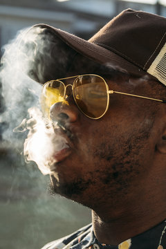Black stylish man wearing sunglasses. He is vaping with an electronic cigarrete outdoor