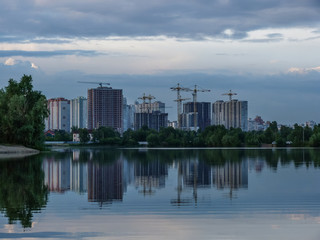 Fototapeta na wymiar new buildings over the lake, the last urban greenery and reflections in the water, unfinished high-rise buildings for monolithic concrete technology, high-rise building cranes, poplar fluff in water