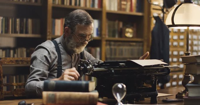Old Caucasian man in glasses typing on the typewriter while sitting in the dark ancient library cabinet.