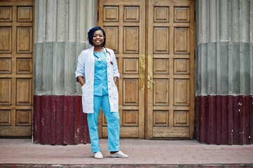 Stylish african american doctor with stethoscope and lab coat posed against door of hospital.