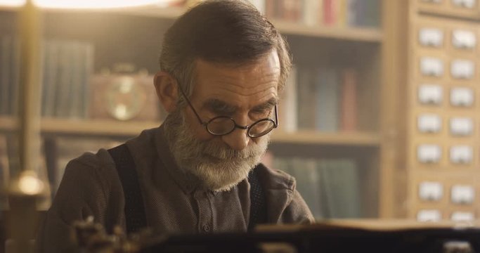 Close up of the old man in glasses starting to type on the typewriter in the ancient atmosphere of the library.