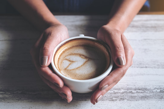Closeup image of woman's hands holding a cup of hot latte coffee on table in cafe