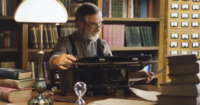 Portrait of the old man in the glasses sitting at the typewriter in the library and thinking.