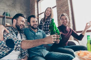 Four attractive, stylish, successful, bearded guys with modern hairstyle clinking bottles with lager, spending holidays together, sitting in living room, laughing and eating snacks, pizza, chips