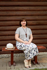 portrait of an elderly woman in a gray linen dress sitting on a wooden bench near a log house in...