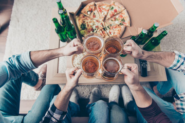 Cropped, top view portrait of hands of company clinking big pints with beer having bottles with lager and snacks on the table, sitting in living room, spending great time