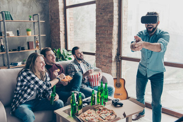 Portrait of amazed attractive bearded guy in vr-headset making gun with fingers like shooting, his happy stylish friends eating chips, popcorn enjoying, laughing at him, sitting in living room