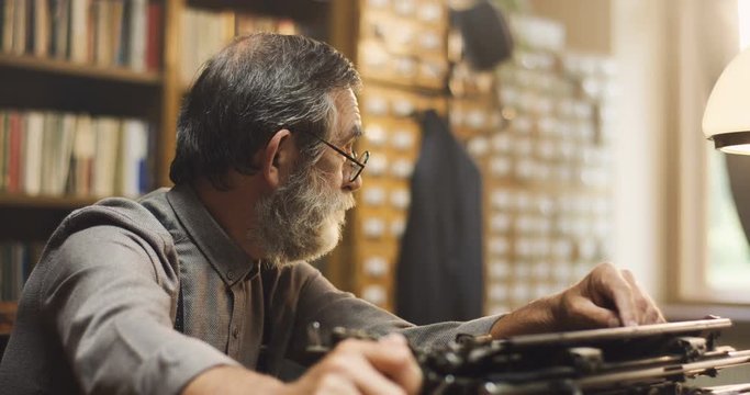 Portrait shot of the old man with beard, gray hair and in glasses thinking while sitting at the typewriter in the library.