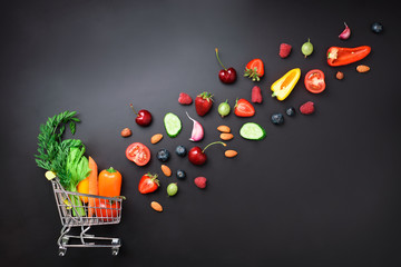 Shopping trolley filled with fresh organic vegetables, fruits and berries on black chalkboard. Top...