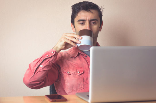 Young man with red shirt drinking coffee while looking at screen of laptop at office desktop. Cellphone on table. Own boss working from home concept