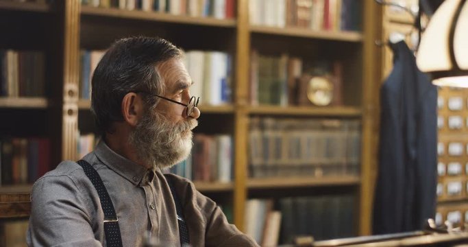 Portrait shot of the old man with beard, gray hair and in glasses thinking about something while sitting in the library. Close up.