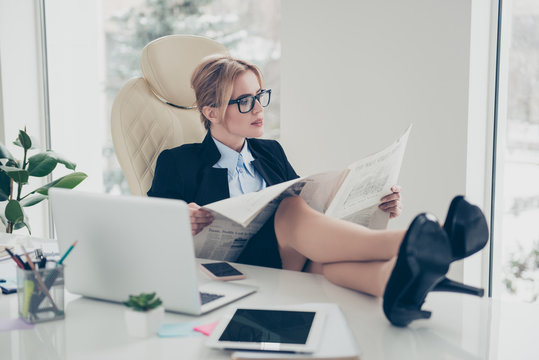 Portrait of experienced stylish woman in eyewear putting legs on table having break reading newspaper sitting in modern workstation