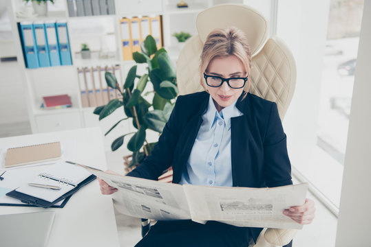 Portrait of clever attractive woman in eyeglasses reading newspaper about policy sitting in modern work place station