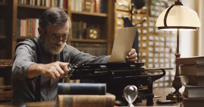 Caucasian old man in glasses putting in paper in the typewriter to start a work in the ancient library.