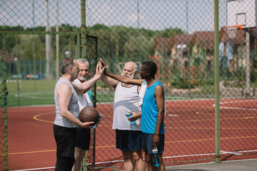 group of multiracial old sportsmen with basketball ball giving high five to each other on playground