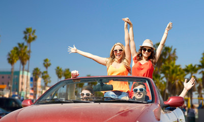 leisure, road trip, travel and people concept - happy friends driving in convertible car at country and waving hands over venice beach background in california - obrazy, fototapety, plakaty