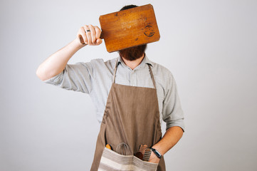 Young chef bearded man covers his face with a wooden cutting board. Man wearing a shirt and an...