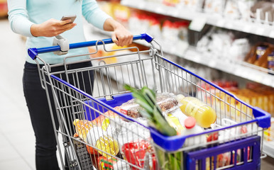 consumerism, technology and people concept - woman with smartphone and shopping cart or trolley...