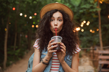 Photo of lovely summer woman 18-20 wearing straw hat, looking at you while drinking beverage from plastic cup during rest in green park with colorful lamps background