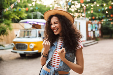 Photo of cheerful happy woman 18-20 with curly brown hair, clenching fists and expressing delight while walk through hipster spot or modern park with colorful lamps background