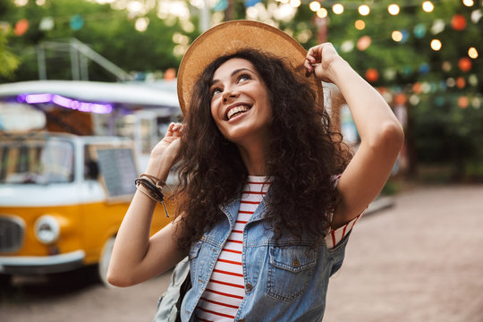 Photo of stylish caucasian woman 18-20 with curly brown hair, laughing and touching her straw hat while rest in hipster spot or modern park with colorful lamps background
