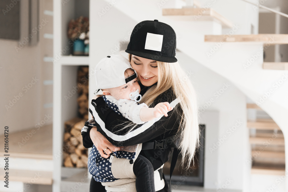 Poster Stylish young mom keeps in ergo backpack of her cute year-old baby at home in a beautiful interior Convenience for mom.