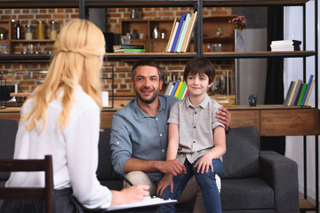 rear view of female counselor writing in clipboard while father and son sitting on therapy session