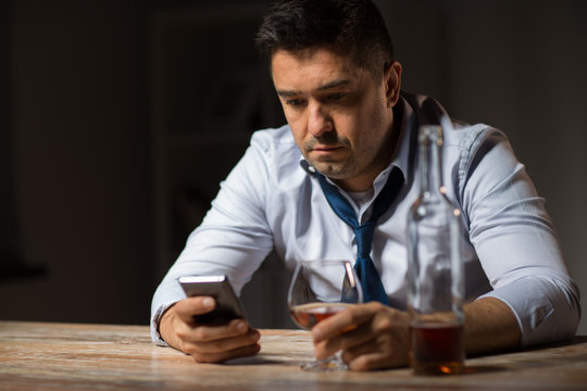 alcoholism, alcohol addiction and technology concept - male alcoholic with smartphone and glass of brandy at night