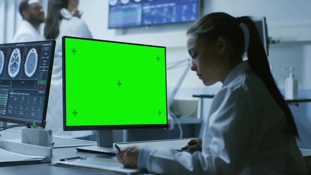 Female Medical Research Scientist Working with Brain Scans on Her Personal Computer Showing Green Mock-up Screen, Writing Down Data in a Clipboard.  Shot on RED EPIC-W 8K Helium Cinema Camera.